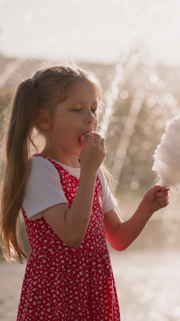 Little boy looks at cotton candy pieces while girl in polka dot sundress eats dessert near fountain with high jets in park slow motion. Sugar snack during walk