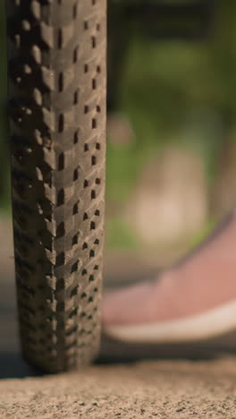 primer plano de las piernas de alguien con zapatillas rosadas pateando un neumático de bicicleta para comprobar la presión del aire, la escena captura la vegetación exuberante y los árboles en el fondo borroso