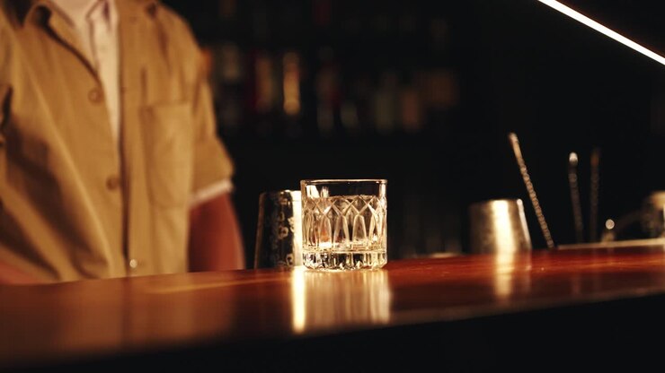 Un barman professionista usa le pinze per posizionare un cubo di ghiaccio trasparente scolpito a mano in un bicchiere rocks di cristallo e lo fa girare per raffreddare il recipiente in un bar di lusso con luci soffuse.