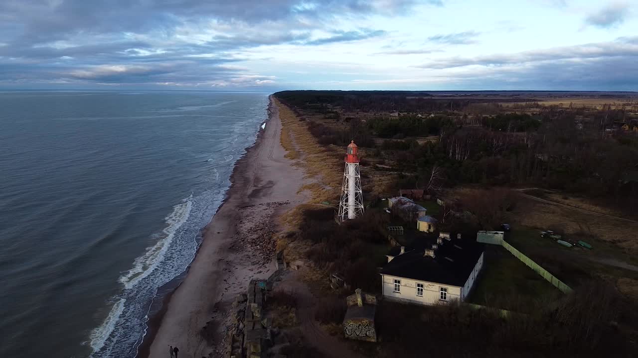 Vista aerea di un faro in acciaio verniciato di bianco situato a Pape, Lettonia, sulla costa del Mar Baltico, in una giornata nuvolosa, con una ripresa grandangolare del drone in movimento all'indietro