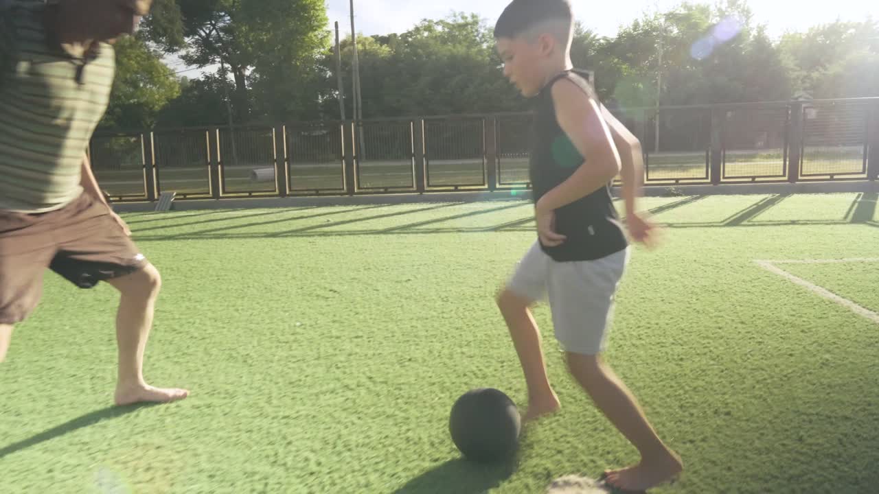 Loving father playing soccer with young son, sharing joyful moment on synthetic grass field during bright summer day