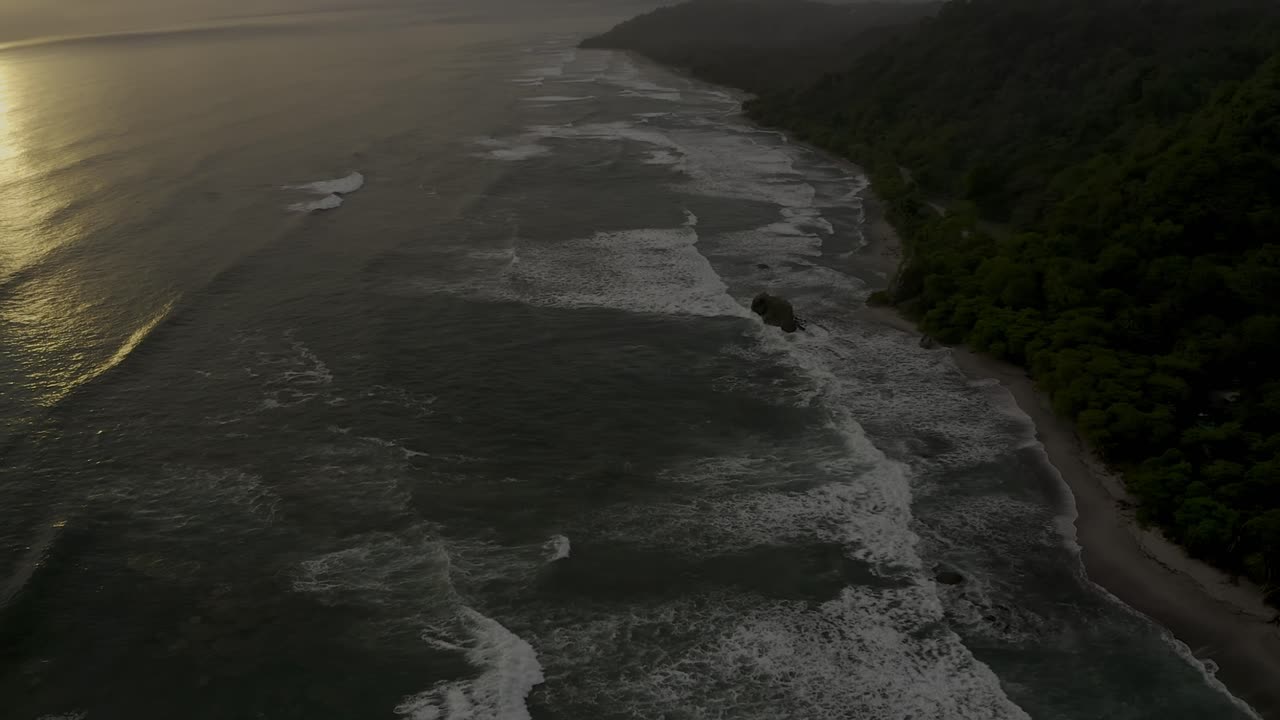 An aerial view captures the serene sunset over Santa Teresa Coast in Costa Rica, with golden waves shimmering under a warm, partly cloudy sky