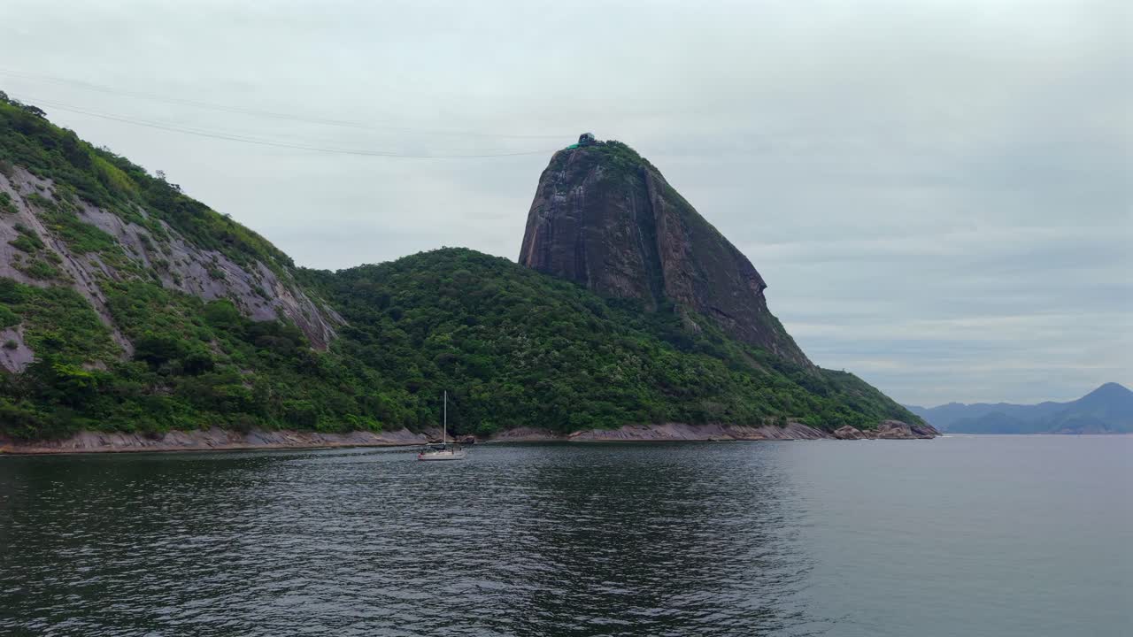Ripresa cinematografica che mostra l'iconica vetta di granito del Pan di Zucchero sulla Baia di Guanabara che si erge dall'Oceano Atlantico con lussureggianti colline verdi e acque blu calme in un paesaggio tropicale brasiliano.