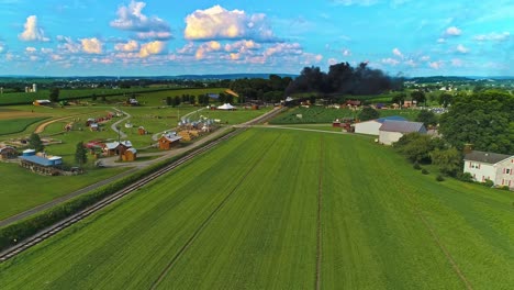 Una-Vista-Aérea-De-Las-Tierras-Agrícolas-Amish-Con-Una-Sola-Vía-Férrea-Y-Un-Tren-De-Pasajeros-A-Vapor-Acercándose