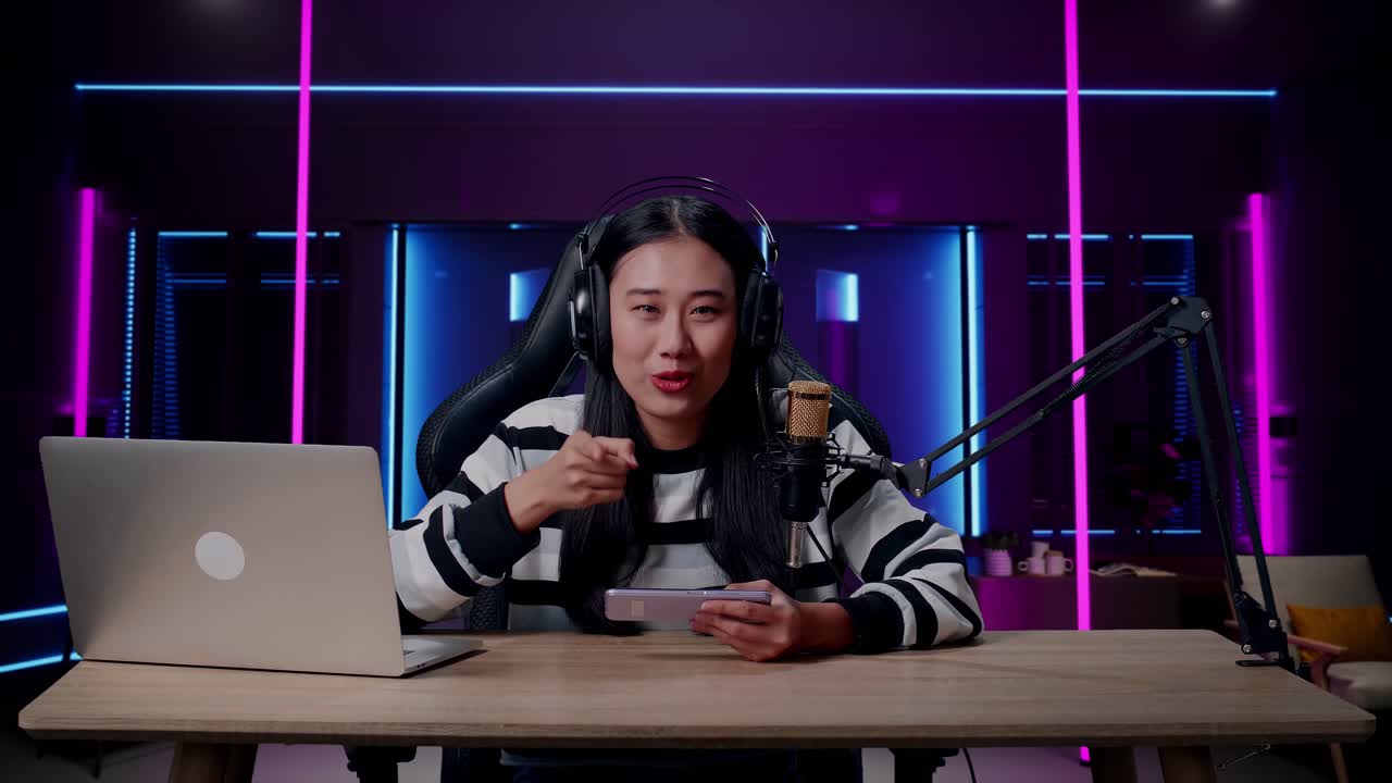 헤드폰과 컴퓨터를 들고 미소 짓는 아시아 여성 게이머가 손을 흔들고 지표손가락을 가리키며 레트로 네온 조명으로 조명된 방에서 휴대 전화 게임을 하고 있습니다.