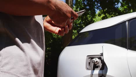 sección media de un hombre mayor afroamericano cargando un coche eléctrico y usando un teléfono inteligente en un día soleado