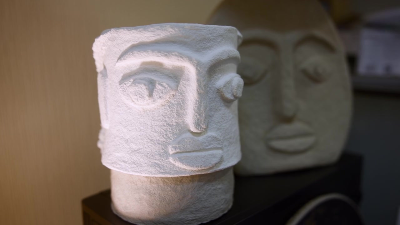 Primo piano di busto scultoreo in gesso con superficie testurizzata, illuminazione da studio calda e morbida, secondo busto sfocato. Media
