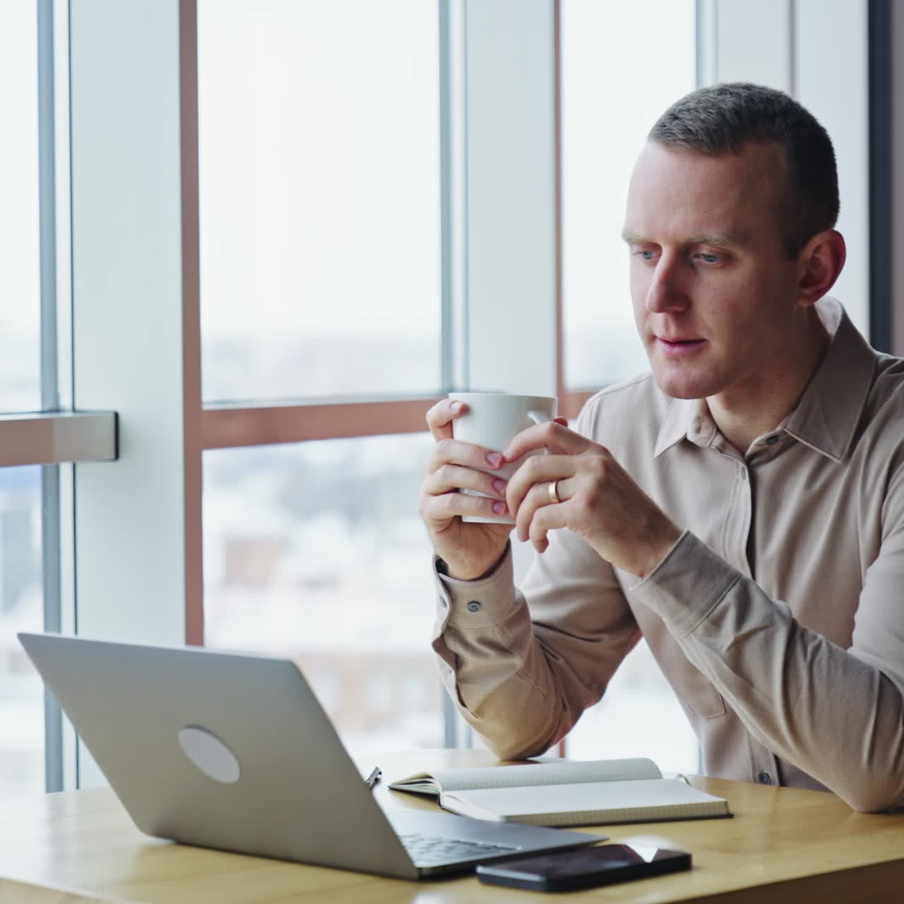 Calm relaxed man sit in front of his computer with a cup in hands. Thoughtful man looks into window and then straight ahead