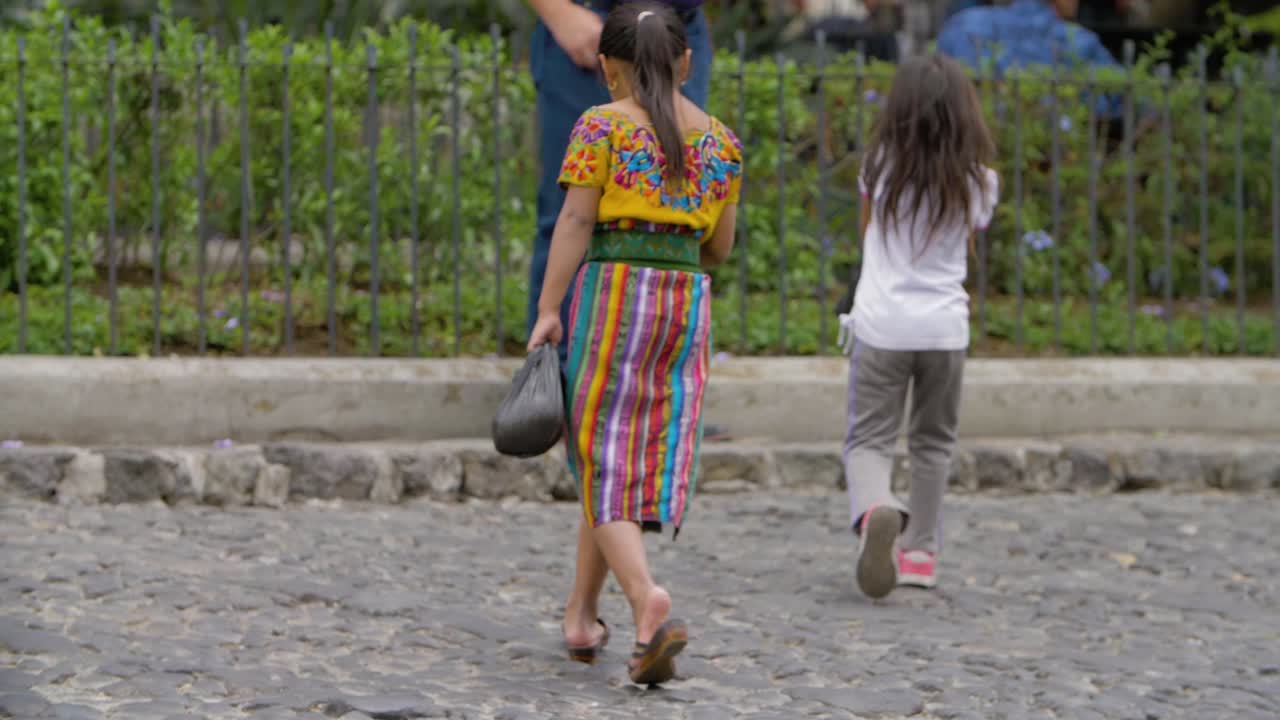 Antigua Guatemala, Sacatepequez pair of young girls cross a cobblestone street as one of them wears a traditional native indigenous dress - Handheld slow motion