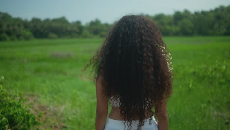 a brunette woman with long wavy hair, dressed in white, stands with her back facing a green field, flowers in her hair