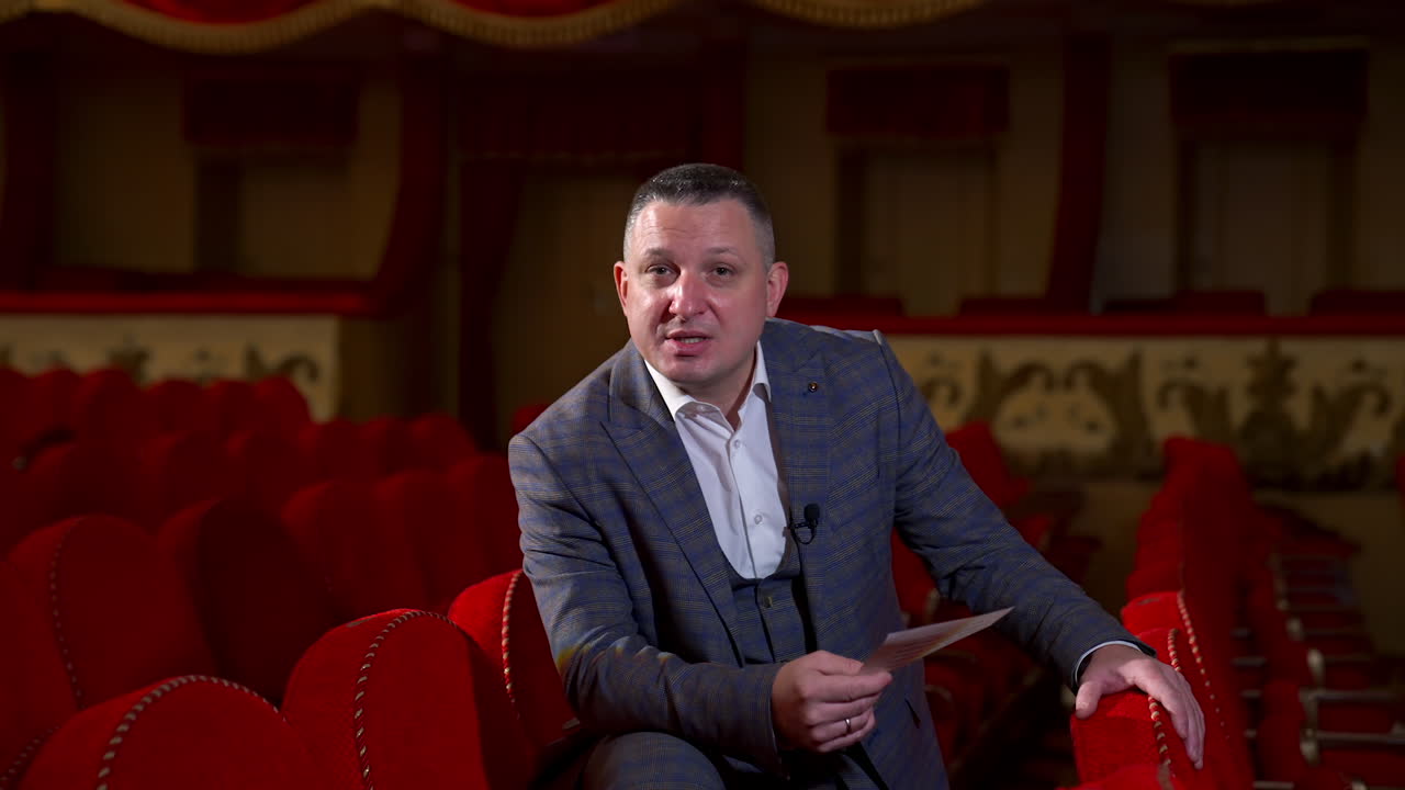 Host announces future theater season. Actor is sitting in empty theater on red velvet chair. Classic theater interior in red colors.