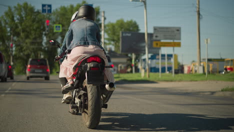 two women riding a power bike slowly approach a traffic light while cars wait for the green light, the rider's friend holds on from behind, a blur of buildings, signs, and cars is seen around