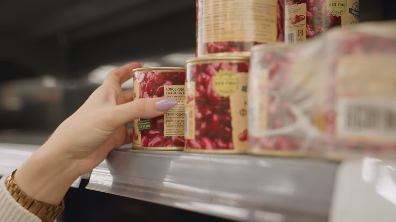 Close up view of female hand with colorful manicure selecting can of red kidney beans from supermarket shelf highlighting vibrant nails and grocery shopping selection in busy retail aisle