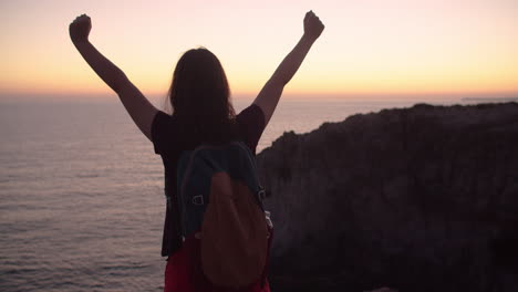 rear view of young girl raising hands in achievement at dawn on top of cliff, slow motion