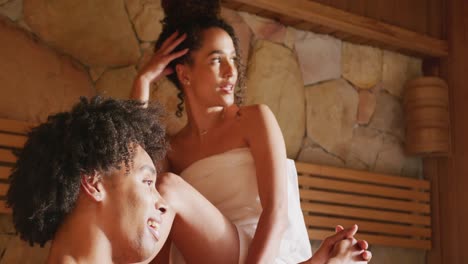 video de una feliz pareja diversa usando toallas relajándose y hablando en la sala de sauna en un spa de salud