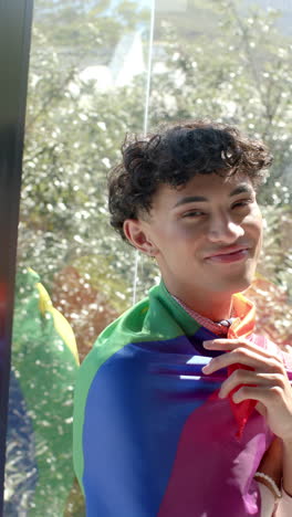 vertical video: smiling person wrapped in rainbow flag standing by window, embracing identity