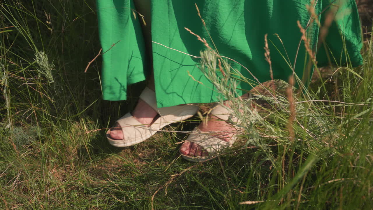 Close up of woman legs in green dress and white sandals stepping carefully through tall summer grass down hillside path under bright sunlight