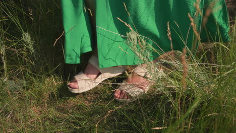 close up of woman legs in green dress and white sandals stepping carefully through tall summer grass down hillside path under bright sunlight
