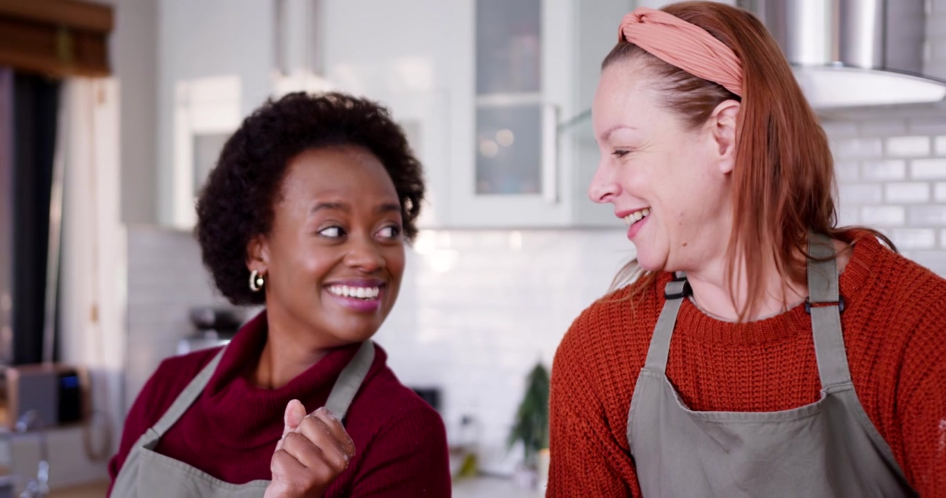 Due donne di diverse etnie, con grembiuli grigi, ridono mentre cucinano al bancone; la donna a sinistra scherza, provocando sorrisi.