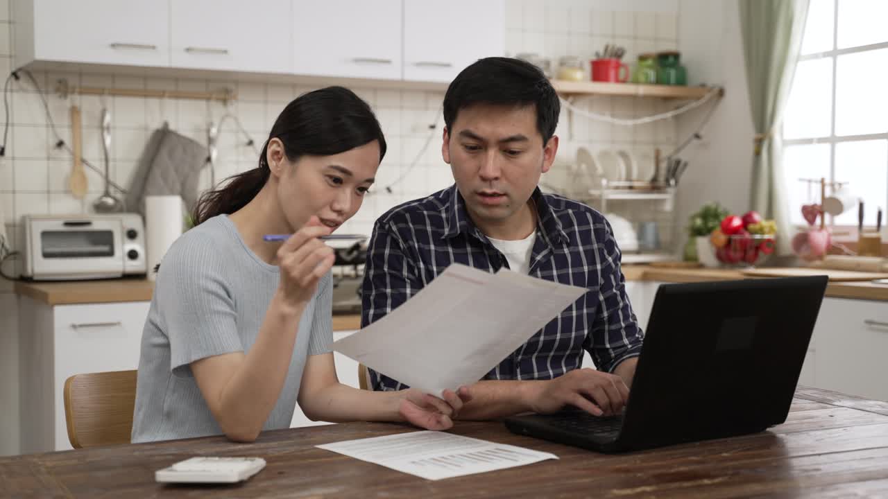 Asian married couple discussing financial plans for the tax season in the dining room at home. they pointing at computer and looking at bills