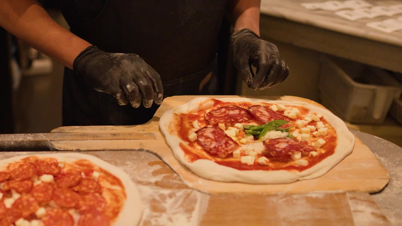 Primo piano di uno chef con guanti neri che assembla una pizza con salame piccante su una pala di legno nella cucina di un ristorante.