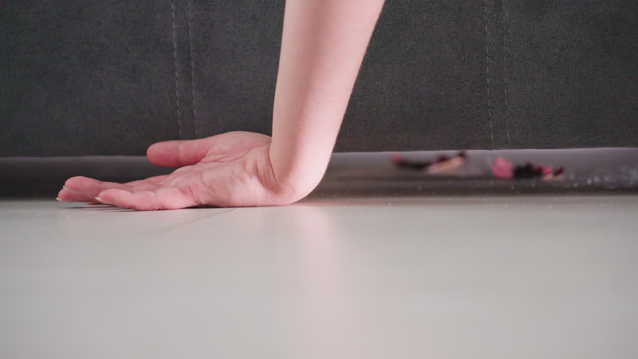 Close up of hand reaching under dark couch touching floor surface near scattered debris and small objects, symbolizing search, cleaning routine, or tidying home environment