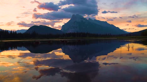 vivid sunset colors fill sky above mountain as still lake mirrors dramatic clouds and rugged peaks in banff national park. tranquil atmosphere enhanced by warm glow and natural reflections