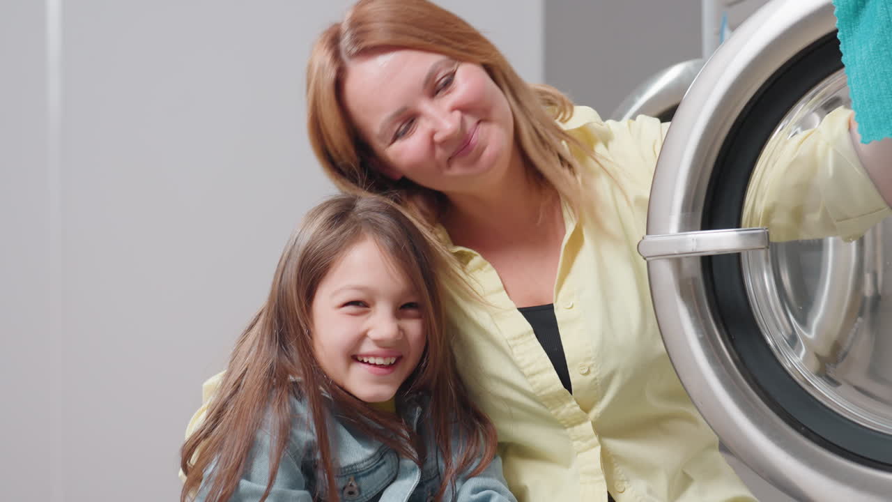 Elegant mother with daughter rest and play beside washer after cleaning, sharing smiles on break inside laundromat, child in denim jacket leaning in hug, stainless door open, family bonding moment