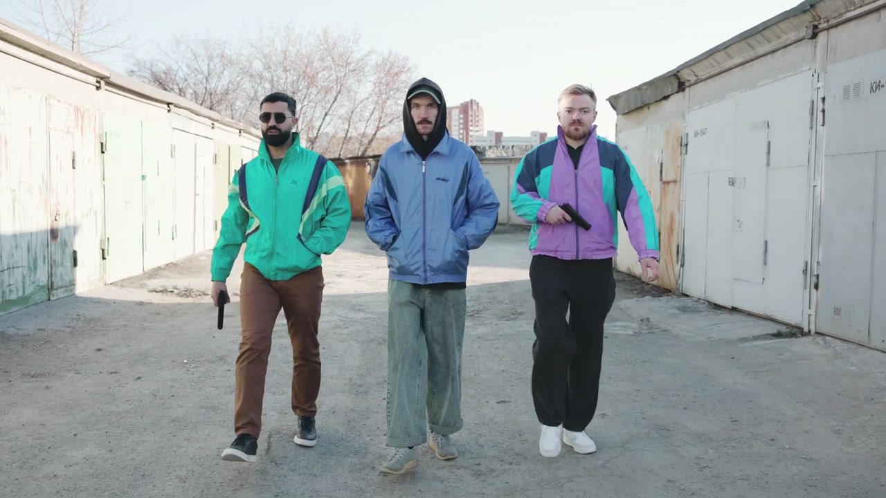 Trois hommes marchant dans une ruelle de garage industriel, vêtus de coupe-vent rétro, les mains dans les poches et d'un pas régulier, par temps froid.