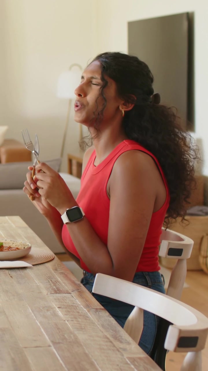 Video verticale: Guardando in basso, una donna in maglietta rossa e jeans prende le posate al tavolo di casa per mangiare.