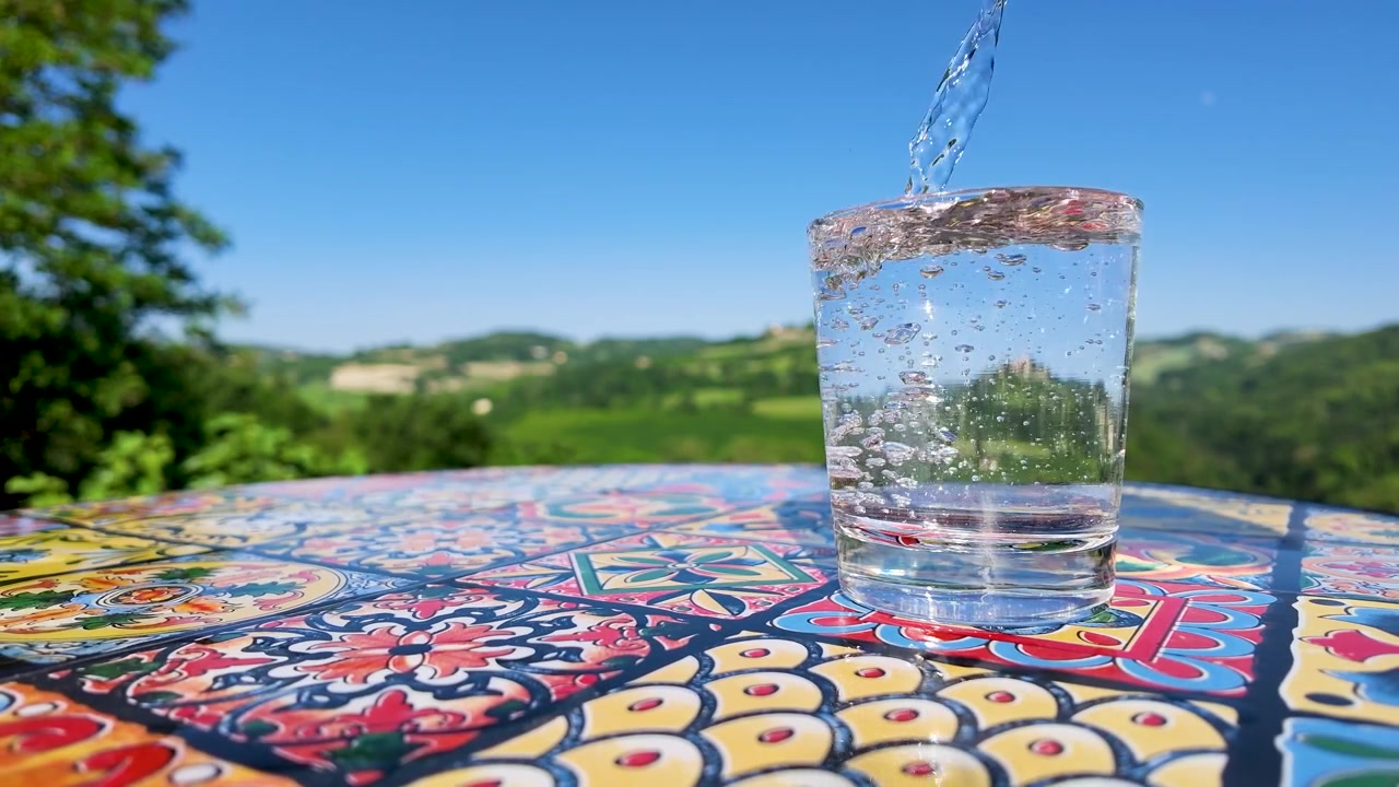 Acqua cristallina e scintillante riempie un bicchiere su un tavolo a mosaico, con uno sfondo di paesaggio toscano.