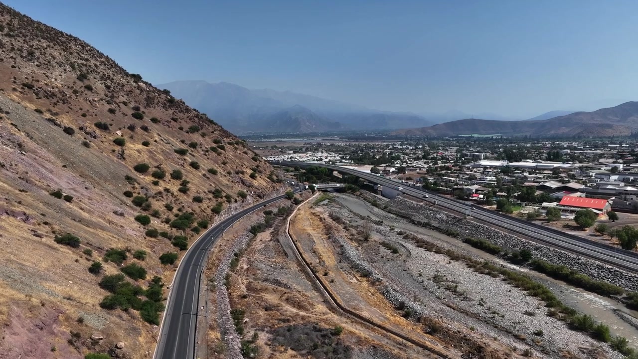Volo in avanti lungo una strada di montagna tortuosa che costeggia un fianco arido della montagna sopra un letto di fiume asciutto, rivelando la città di Los Andes che si estende attraverso la valle dell'Aconcagua con le cime andine sullo sfondo. Los Andes, Cile.