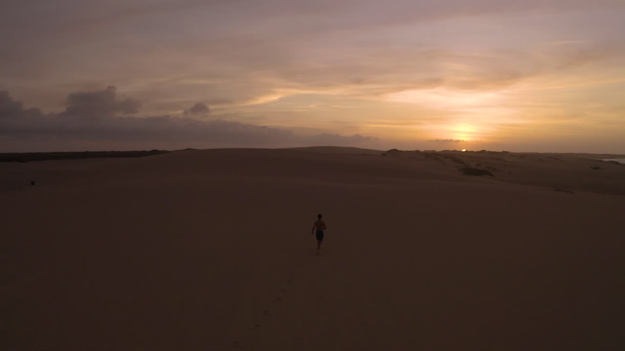 toma aérea de un joven corriendo hacia la puesta de sol en un desierto