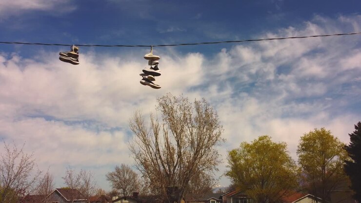 Slow drone pan across telephone wire with tennis shoes trainers on it with blue sky and white clouds, spring trees and rural suburban neighborhood houses in background