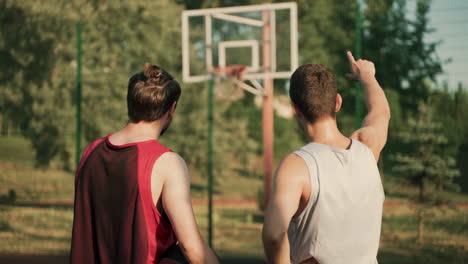 back view of two handsome male basketball players looking at basket and talking about game tactics in a blurred outdoor basketball court in a sunny day