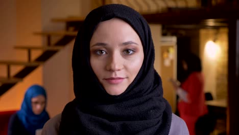 closeup portrait of young beautiful arabian female office worker in black hijab looking straight at camera and smiling confidently being on the workplace