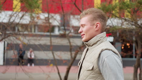 young man walking along dry autumn trees as wind blows gently, wearing light jacket with mall in background and people standing nearby
