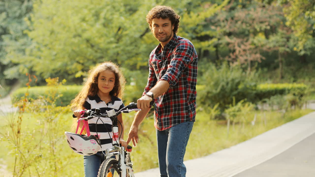 Portrait of a little girl and her dad standing near the bike. They look at each other and then - into the camera and smile. Blurred background