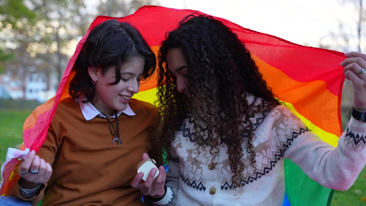 Young smiling female couple covered with a pride flag, lovingly looking at each other in a park