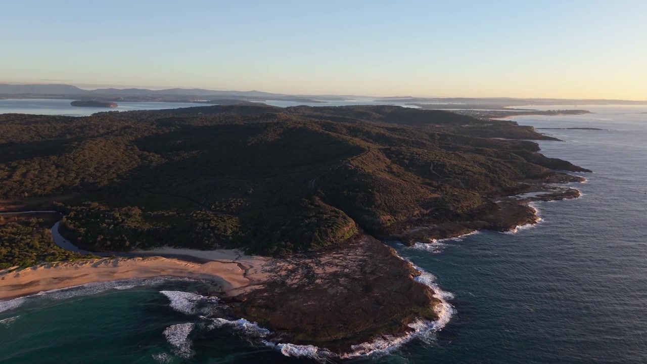 Promontorio coperto da vegetazione che incontra la costa rocciosa e l'oceano blu all'alba nel Nuovo Galles del Sud, Australia, ripresa aerea a media distanza, sfondo naturale