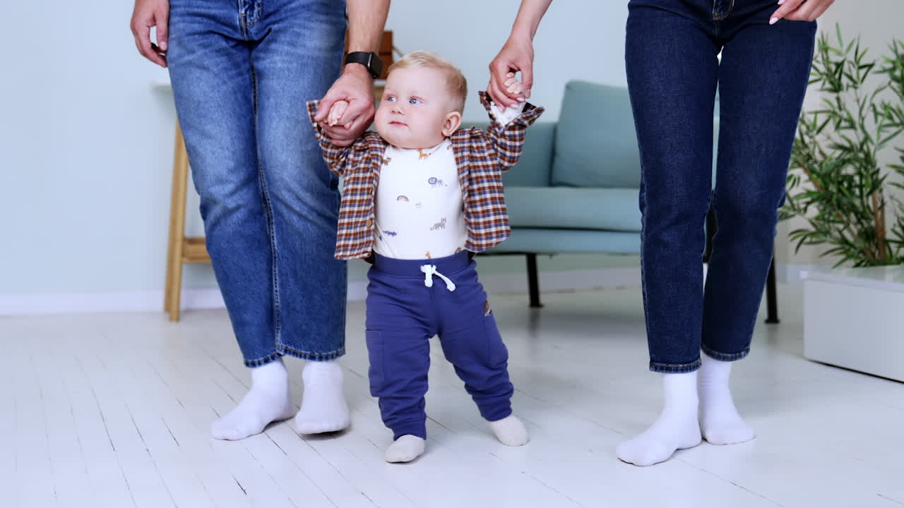 Unrecognized parents wearing jeans hold their toddler son. Lovely cute baby boy learning to walk.