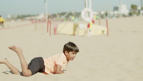 Vista-Frontal-De-Un-Niño-Emocionado-Pateando-Una-Pelota-Y-Cayendo-Sobre-La-Arena.