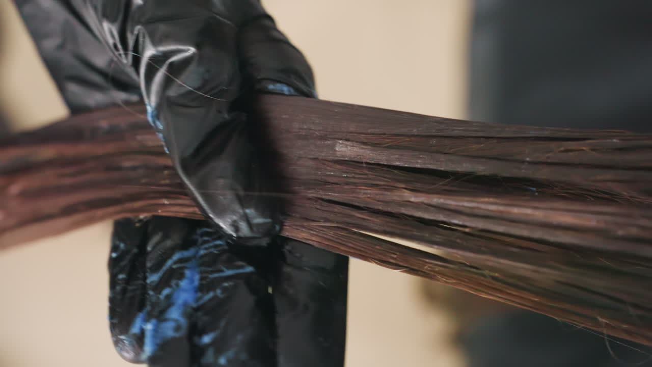 젖은 머리카락에 염색약을 클로즈업하여 바르는 모습이에요. 장갑을 낀 손으로 염색약을 바르고 빗질하여 색소를 입히고, 윤기 있는 결과를 위해 포뮬러 혼합, 코팅 과정, 임상 살롱의 정밀함을 보여줍니다.
