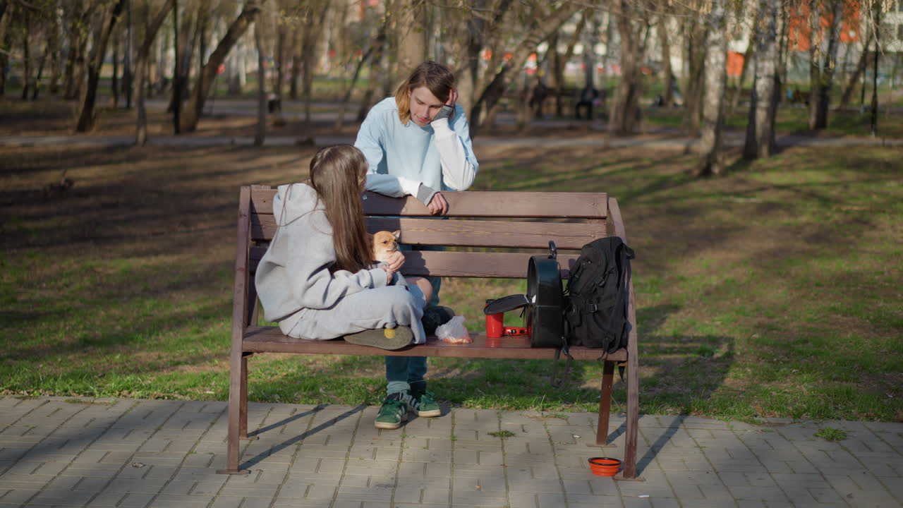 Asiatiske og kaukasiske teenagere slapper af sammen udendørs. Teenagere deler mad og latter under de grønne parktræer. Unge venner med forskellige baggrunde snakker og spiser snacks midt på dagen i parken.