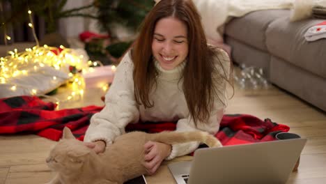 Happy-brunette-girl-in-a-White-sweater-lies-on-the-floor-on-a-Red-blanket-near-her-cream-cat,-strokes-it-and-watches-a-movie-on-a-gray-laptop-in-a-cozy-room-decorated-in-Christmas-style