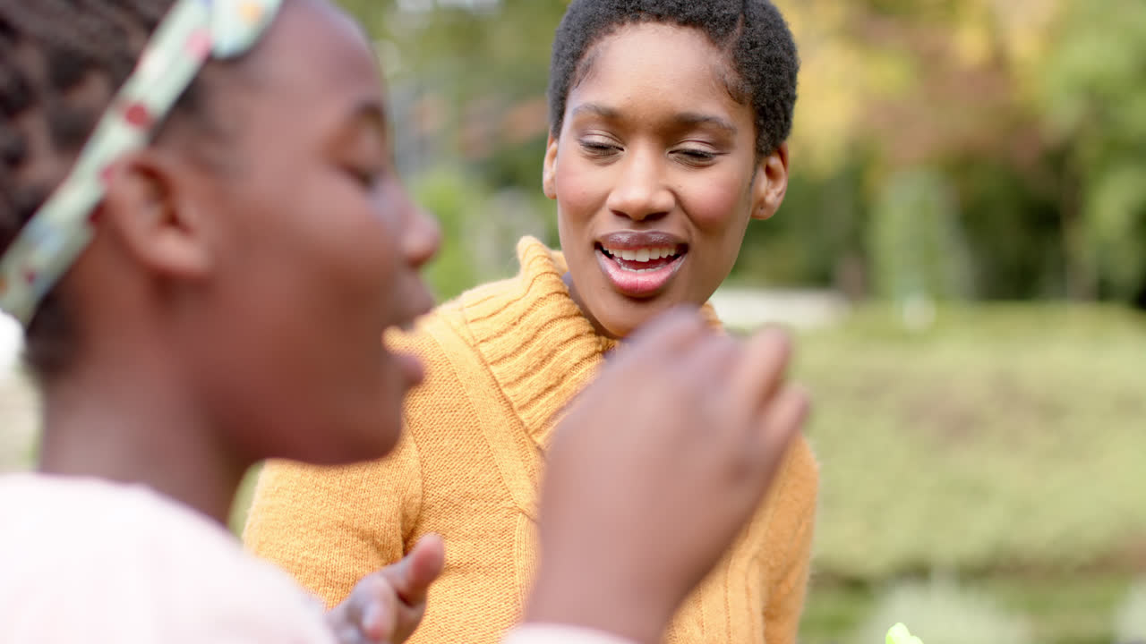 madre y hija afroamericanas felices soplando burbujas en el jardín soleado