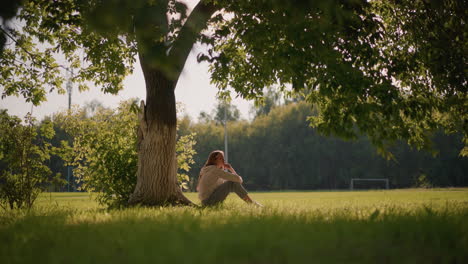 woman sits thoughtfully on grassy ground with hand on chin under tree shade, sunlight filters through leaves, in background, blurred figure moves across open field with distant goalpost
