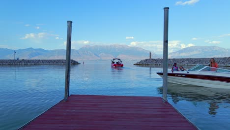 Ein-Familienmotorboot-Dreht-Sich-Vor-Einem-Hölzernen-Ponton,-Bereit-Zum-Docken,-Mit-Einem-Leuchtend-Roten-Motorboot,-Das-Im-Hintergrund-Den-Hafen-Von-Saratoga-Springs-Verlässt,-Umgeben-Von-Einer-Sonnenbeschienenen-Bergkette