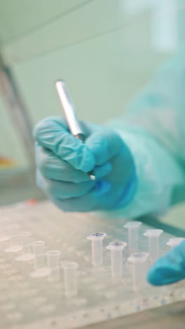 laboratory worker writes with a pen on test tubes. hands in blue protective gloves writing on plastic vials on the table indoors. close-up. vertical video