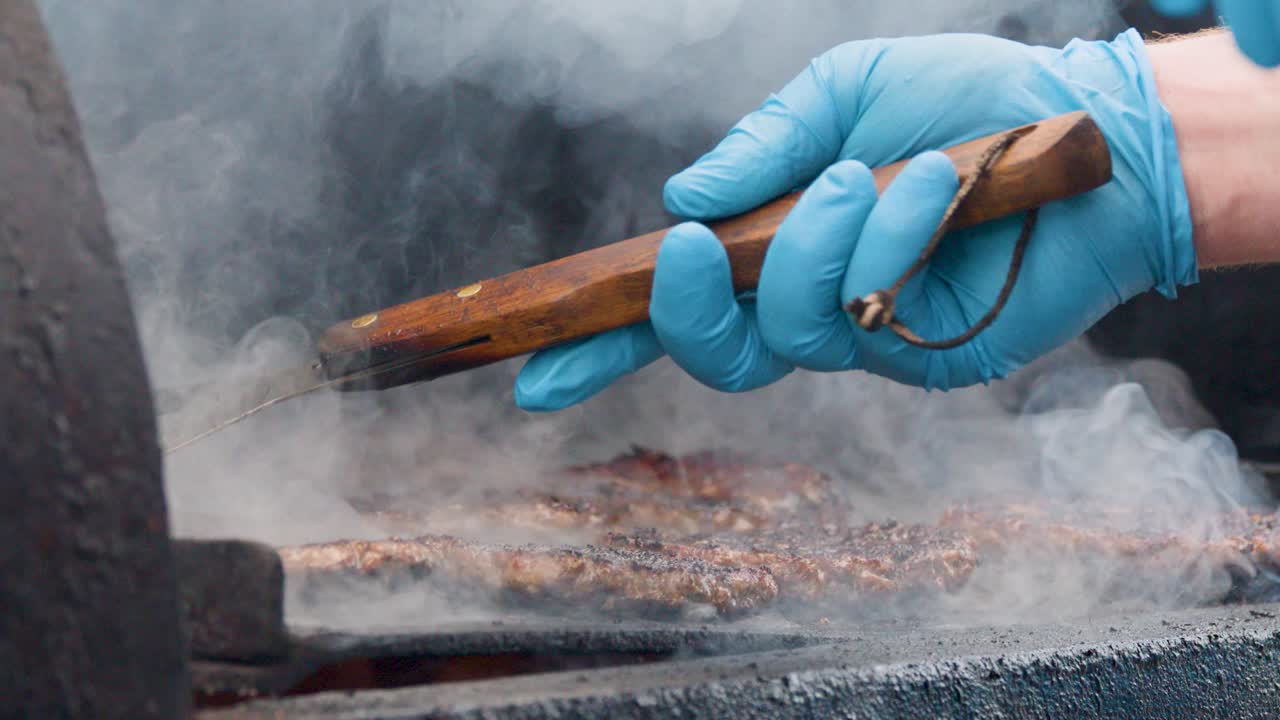 Blue-gloved hand uses spatula to flip burgers on smoky barbecue grill, close-up shot